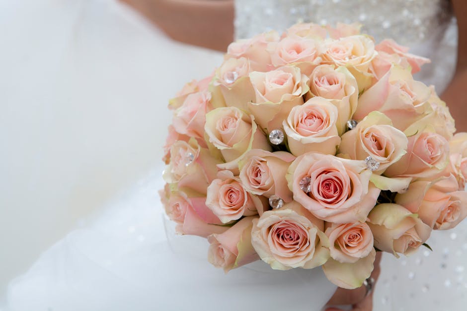 A close-up of a bride holding a vibrant bouquet of fresh wedding flowers, including white roses, blue hydrangeas, and white dahlias, complemented by lush green foliage. The bouquet is arranged in a ro