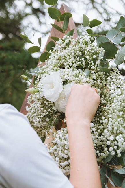 A bride holds a bouquet arrangement featuring cream and ivory roses, along with small white daisies, blue thistles, and eucalyptus leaves, arranged in a natural, slightly asymmetrical style. The bouqu