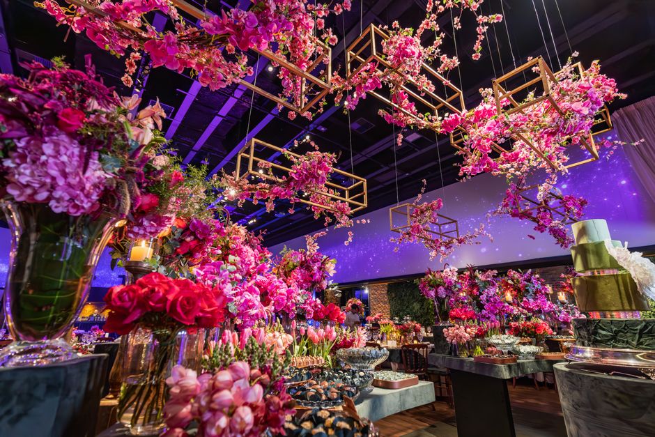 A long banquet table set for a formal occasion, decorated with elaborate floral arrangements featuring pink, white, and pale blue flowers, including roses and hydrangeas, in tall, ornate gold stands. 